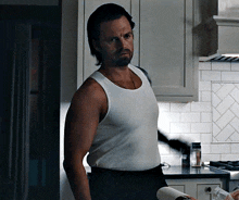 a man in a white tank top is standing in a kitchen holding a towel .