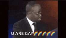 a man in a suit and tie is saying `` u are gay '' with rainbows in the background .