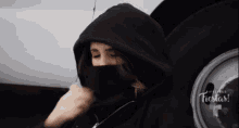 a woman wearing a black hoodie is sitting next to a white car