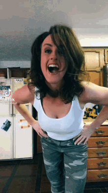 a woman wearing a white tank top and camo pants is standing in front of a refrigerator with a tik tok sticker on it