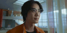 a young man wearing glasses and an orange jacket is standing in a room .
