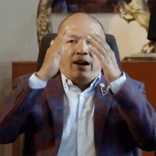 a bald man in a suit is covering his eyes with his hands while sitting in a chair .
