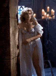 a woman in a long white dress is standing next to a wall with candles .