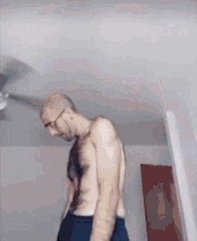 a shirtless man with glasses is standing in a room with a ceiling fan .