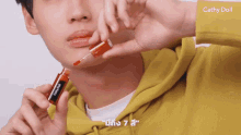 a man wearing a yellow hoodie is holding a bottle of cathy doll lip tint