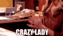 a woman is sitting at a desk with her hands up and the words `` crazy lady '' written on the screen .