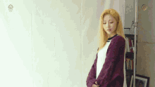 a woman with blonde hair wearing a purple cardigan stands in front of a white wall
