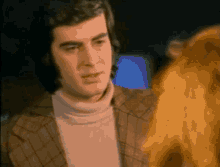 a man in a plaid jacket and a white turtleneck is looking at a woman