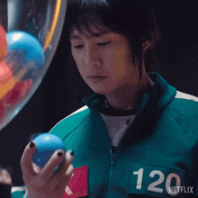 a woman in a green jacket with the number 120 on the back is holding a blue ball .