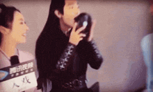 a man in a black jacket is holding a black object in his hand while a woman looks on