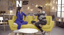 a man and a woman are sitting on a yellow couch in front of a wall with letters ns