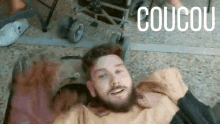 a man with a beard is laying on the floor with a stroller in the background and the word coucou above him .