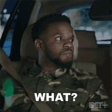 a man in a camo shirt is sitting in the driver 's seat of a car and says " what "