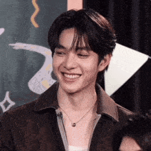 a young man wearing a necklace and a brown jacket is smiling .