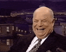 a bald man in a suit and tie is laughing while sitting in front of a city .