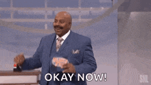 a man in a suit and tie is standing on a stage and saying `` okay now ! ''