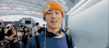 a man wearing an orange beanie and a blue shirt is standing in front of a crowd at an airport