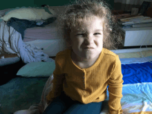 a little girl in a yellow shirt is sitting on a bed making a funny face