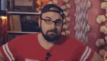 a man with a beard and glasses is wearing a red shirt and a black hat .