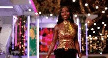 a woman in a gold top and black pants stands in front of lights