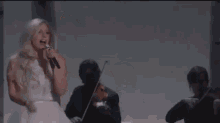 a woman in a white dress is singing into a microphone while a man plays violin in the background .