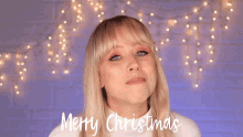 a blonde woman says merry christmas in front of christmas lights