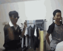 two men are standing in a room with clothes hanging on a rack