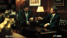two men in suits are sitting at a table with a nbc logo on the bottom