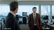 a man in a suit and tie talks to another man in an office with the nbc logo in the background