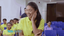 a woman in a yellow shirt is smiling in front of a group of people in a lecture hall .