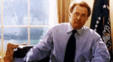 a man in a blue shirt and tie is sitting at a desk in front of an american flag