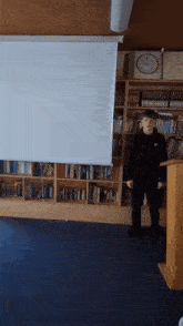 a man standing in front of a projector screen with a clock on the wall behind him