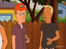 two men are standing next to each other in front of a wooden fence and one is wearing an orange hat