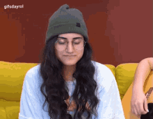 a woman wearing a green beanie and glasses is sitting on a yellow couch ..