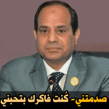 a man in a suit and tie stands in front of a microphone with arabic writing behind him