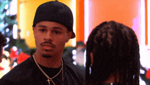 a man wearing a hat and a cross necklace looks at another man with dreadlocks