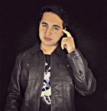 a young man wearing a black leather jacket holds his finger to his forehead while standing in front of a black background