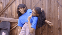a woman in a blue shirt holds another woman on her shoulders in front of a wooden fence