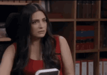 a woman in a red dress is sitting at a desk in front of a bookshelf and looking at the camera .