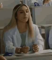 a woman in a lab coat is sitting at a desk in a classroom with other people .
