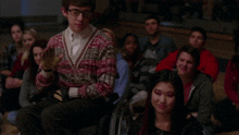 a man in a sweater that says ' sweater ' on it sits in front of a crowd of people