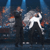 two men singing into microphones on a stage with a picture of a man behind them