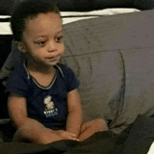 a baby in a blue shirt is sitting on a bed looking at something .