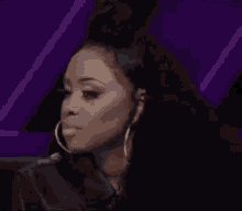 a woman wearing hoop earrings is sitting in front of a purple background and talking on a cell phone .