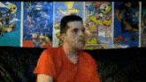 a man in an orange shirt is sitting in front of a wall with posters of cartoon characters