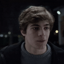 a young man wearing a grey sweater and a black jacket smiles