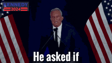 a man in a suit and tie is giving a speech in front of an american flag with a kennedy 2024 sign behind him