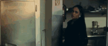 a woman in a black coat is standing in a kitchen next to a refrigerator .