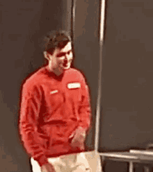 a man wearing a red sweater and white pants is standing in front of a black wall and smiling .