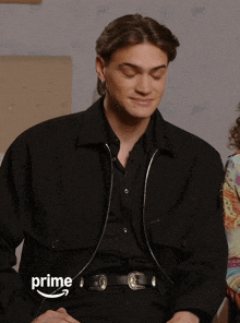 a man wearing a black shirt and a black jacket is sitting in front of an amazon prime logo